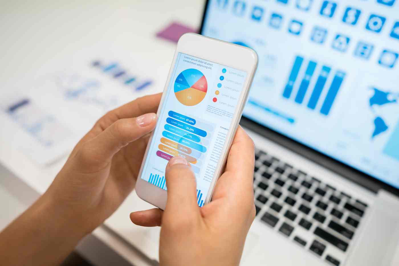 A person's hands holding a smartphone displaying charts and other information in the foreground, with a computer in the background displaying a map and charts.