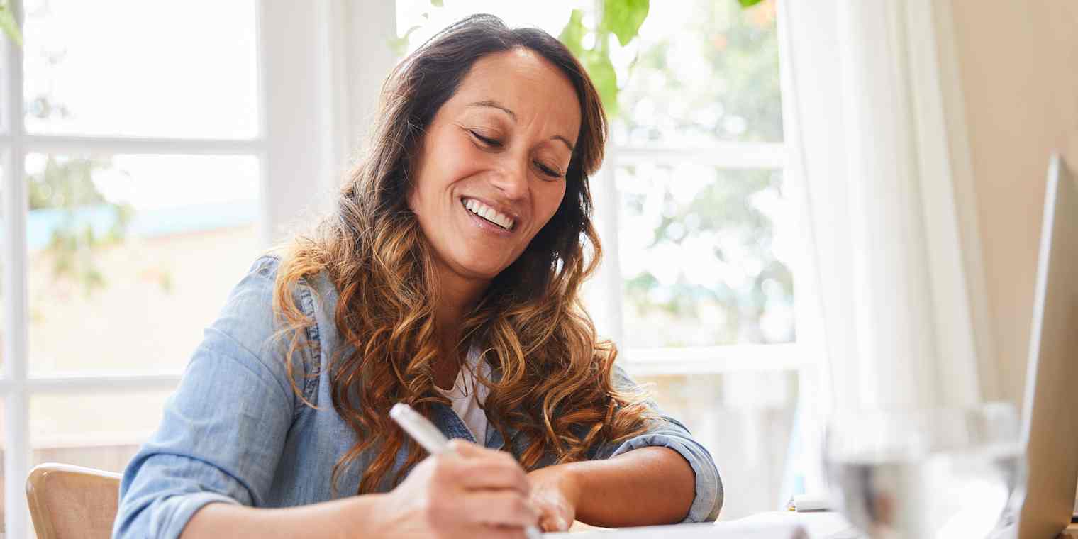 A woman sits at a table working. She has a computer and a notebook in front of her.