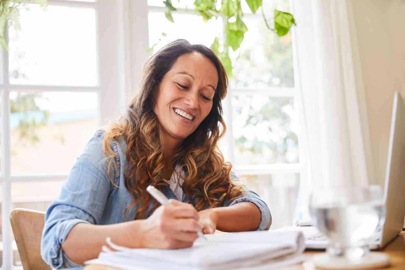 A woman sits at a table working. She has a computer and a notebook in front of her.