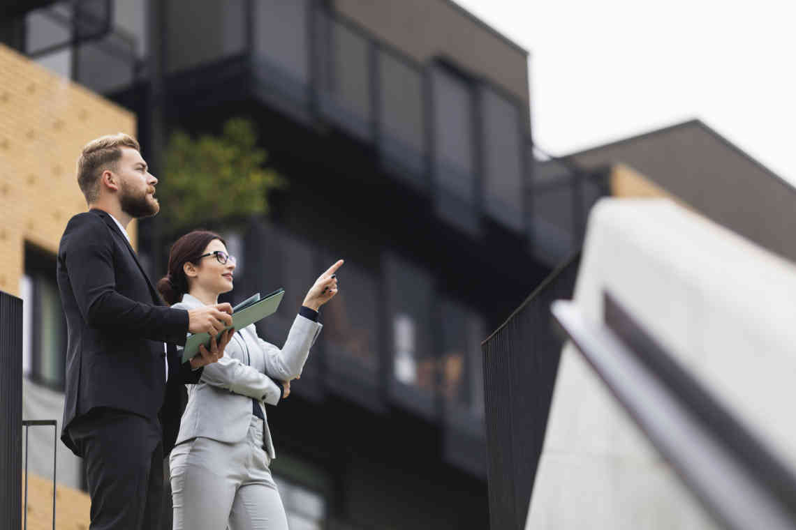 A man and woman stand outside looking at properties. He is carrying a folder and she is pointing up at a building.