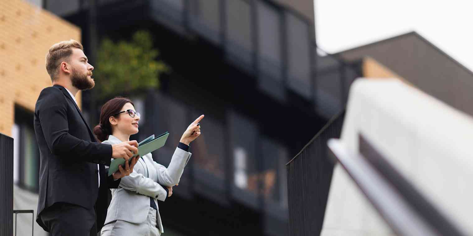 A man and woman stand outside looking at properties. He is carrying a folder and she is pointing up at a building.