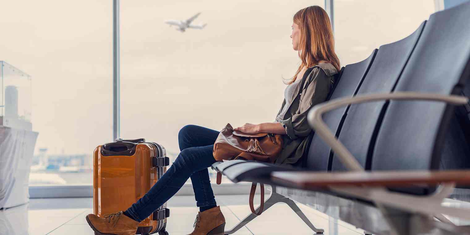 A blonde woman sits at an airline gate with a suitcase on the floor next to her. An airplane is visible through the windows.