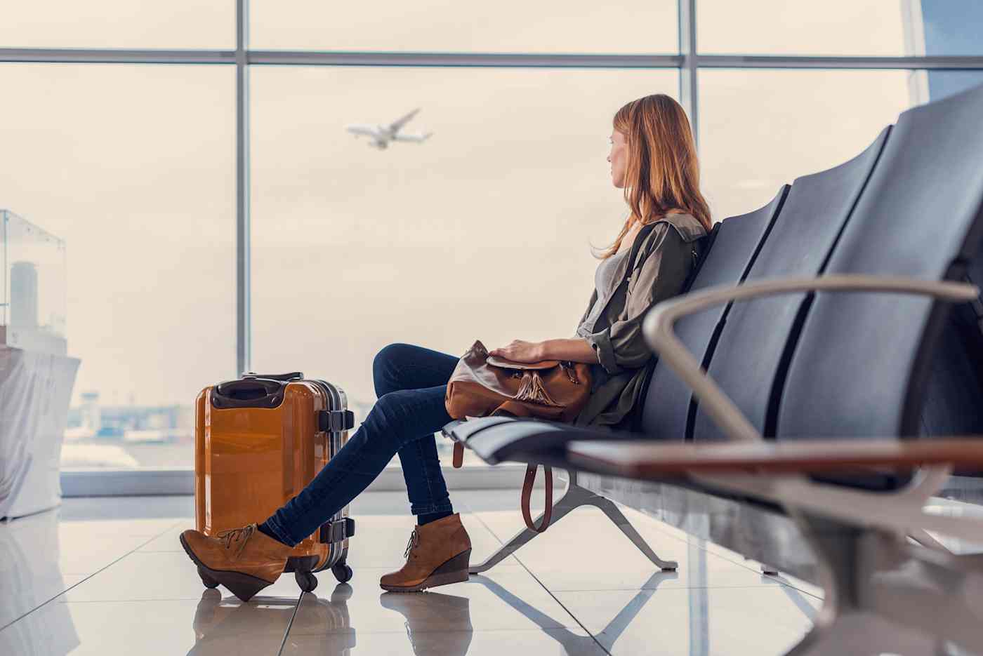 A blonde woman sits at an airline gate with a suitcase on the floor next to her. An airplane is visible through the windows.