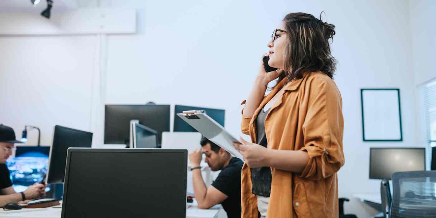 Hero image of a woman talking on a phone holding a clipboard in an office with other people around