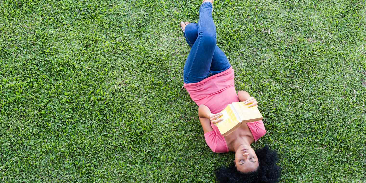 A woman wearing casual clothes lies on her back on a green lawn. She is reading a book.