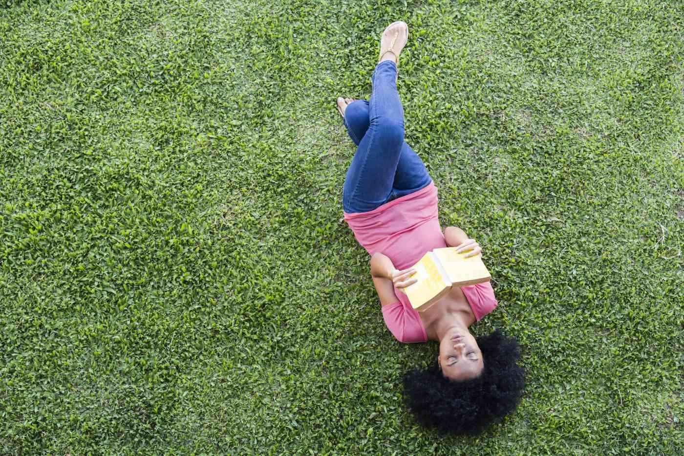 A woman wearing casual clothes lies on her back on a green lawn. She is reading a book.
