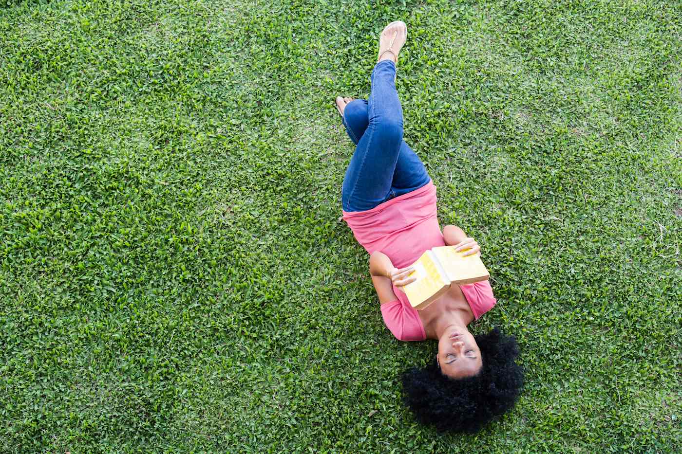 A woman wearing casual clothes lies on her back on a green lawn. She is reading a book.