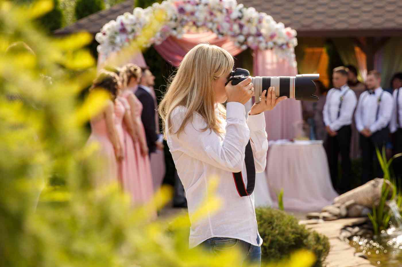 A woman wearing a white shirt holds a camera with a long lens up to her face. Out of focus in the background is a wedding party and a flowered canopy.