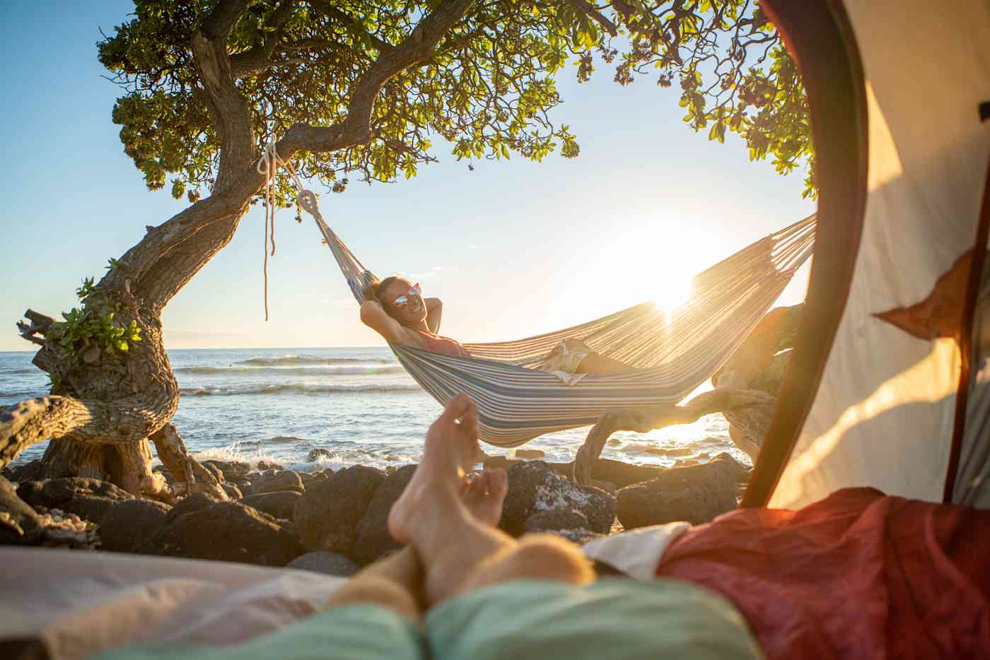 This photo is taken from the perspective of someone lying on the ground inside a tent, looking through the opening. In the foreground, you can see their legs, crossed at the ankle. They are looking at a woman relaxing in a hammock next to water.