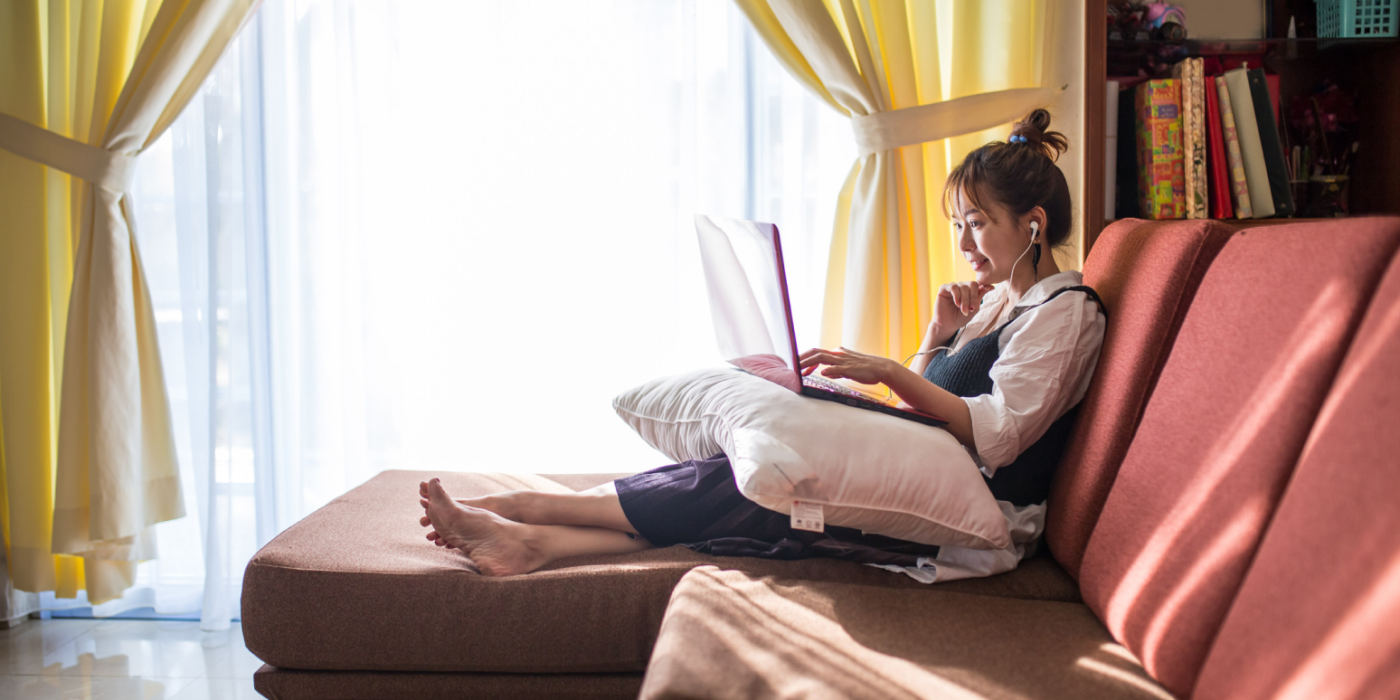 Hero image of a woman sitting on a couch, with a pillow on her lap and a laptop on the pillow, with headphones in