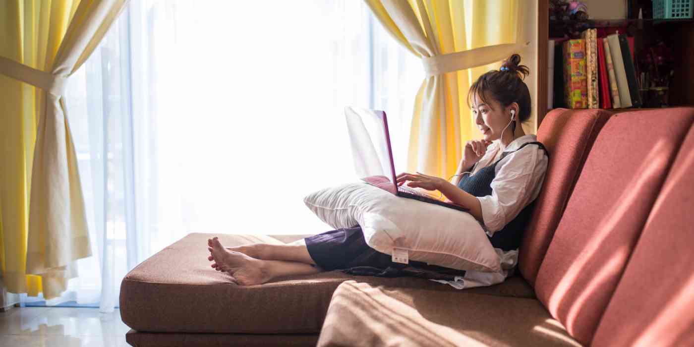 Hero image of a woman sitting on a couch, with a pillow on her lap and a laptop on the pillow, with headphones in