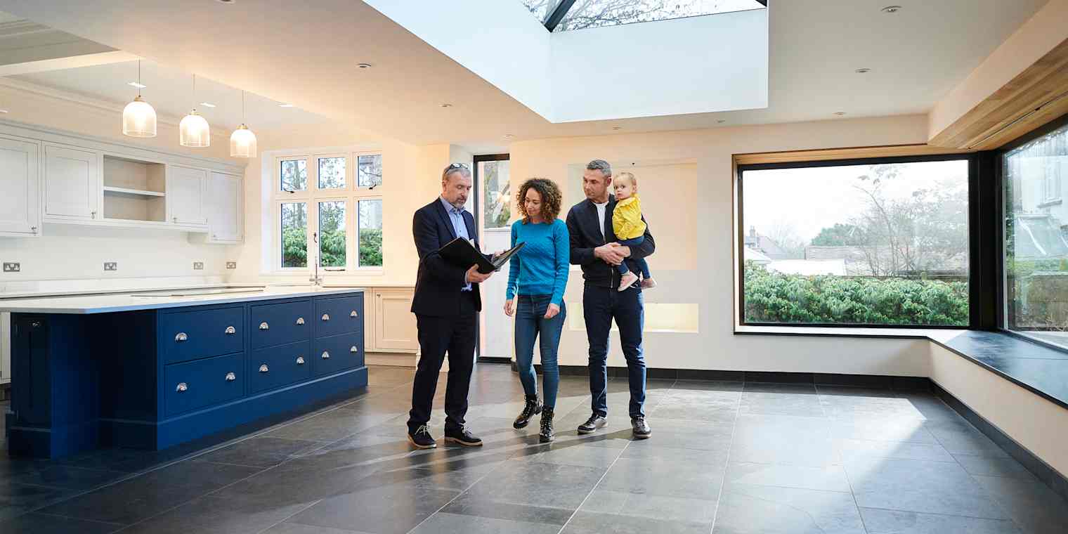 A real estate agent shows a vacant home to a couple with a small child. They are standing in a large kitchen.