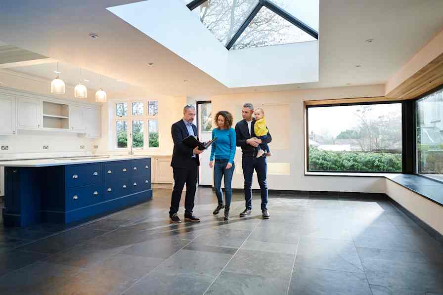 A real estate agent shows a vacant home to a couple with a small child. They are standing in a large kitchen.