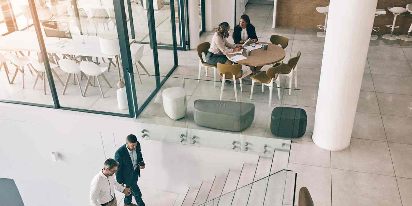 Hero image of the inside of a big, modern office building, with two people walking up the stairs and two people sitting at a table