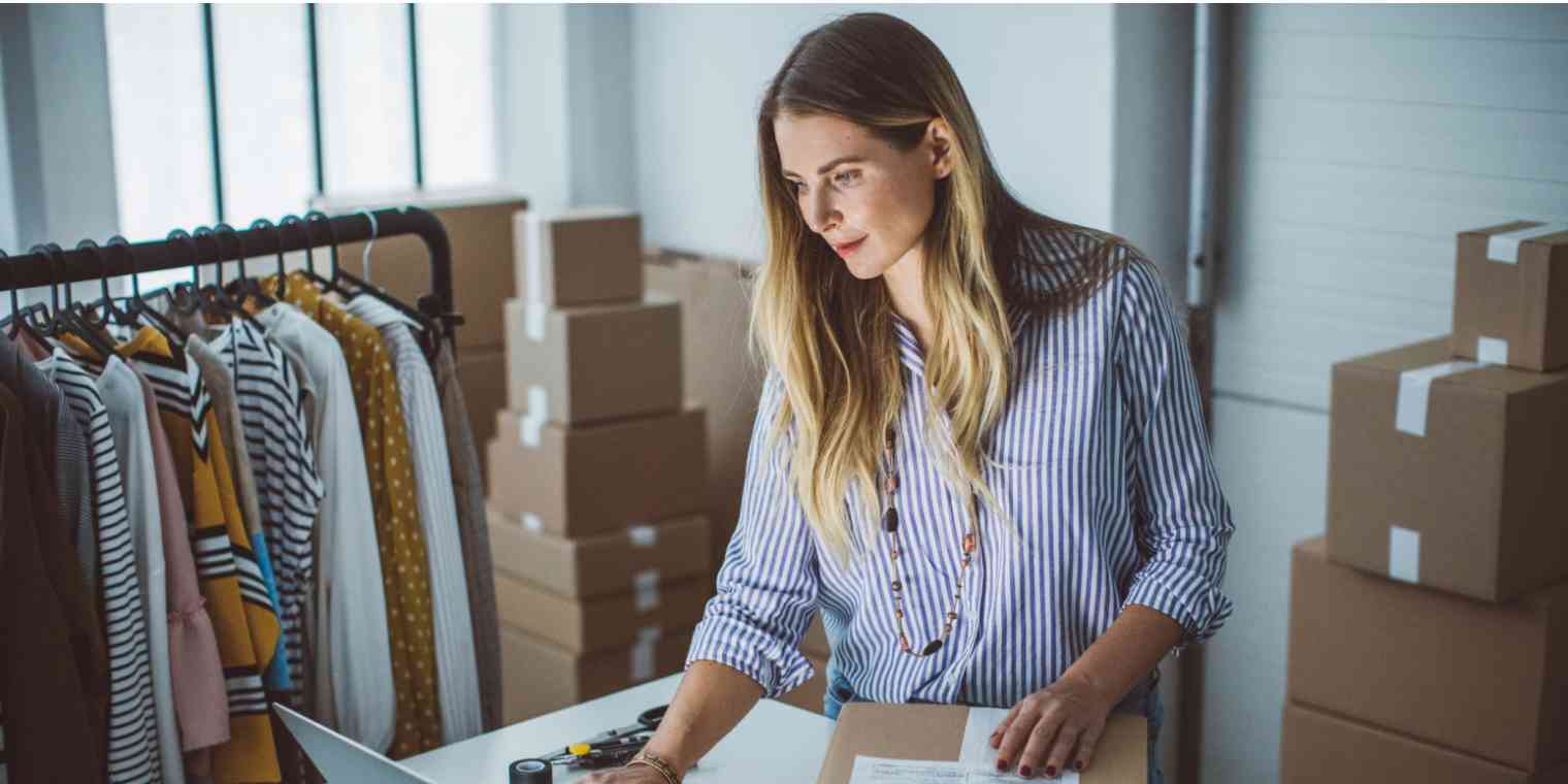 Hero image of a woman in a room of boxes and clothes, looking at a computer