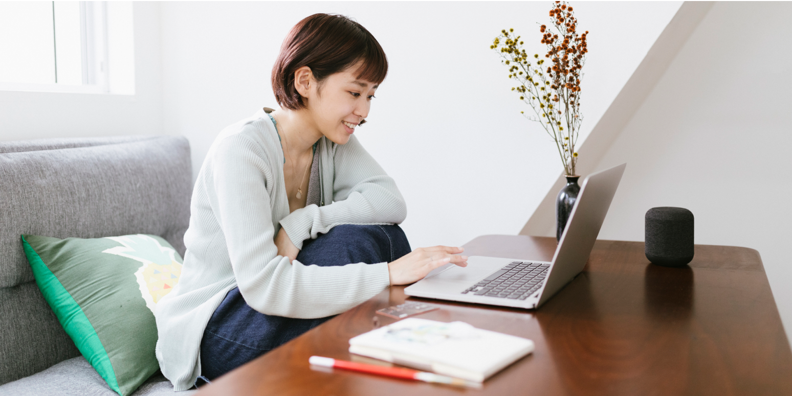 Hero image of a woman on a couch, with a table in front of her, looking at the computer and smiling