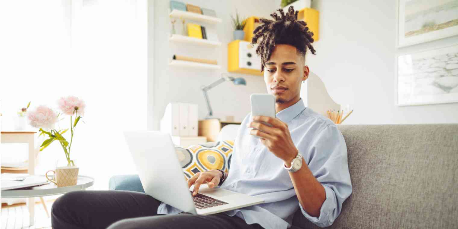 Screenshot of a man on a couch with a computer on his lap, holding and looking at a phone