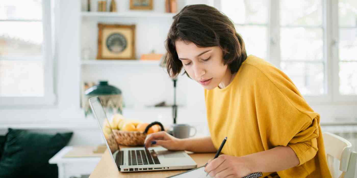 Hero image of a woman in a bright yellow shirt in front of a computer, writing something on a notepad