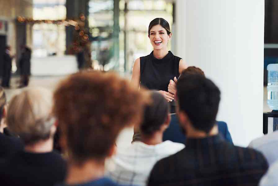 A smiling woman speaks to a group of seated people at an event.