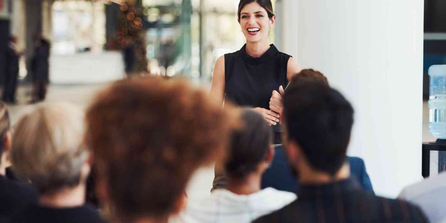 A smiling woman speaks to a group of seated people at an event.