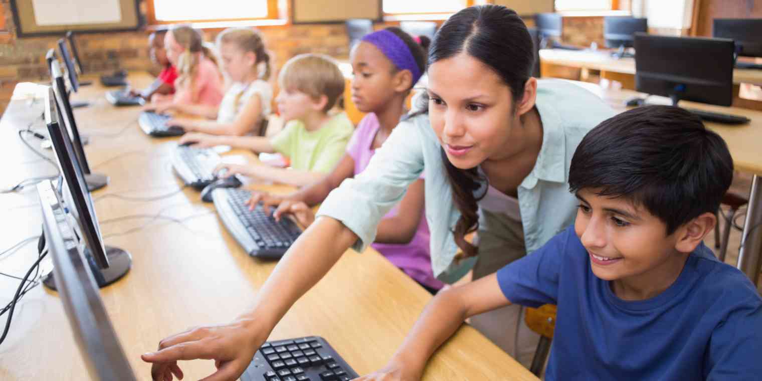 Hero image of a teacher leaning over a computer with a student, in a row of students at computers
