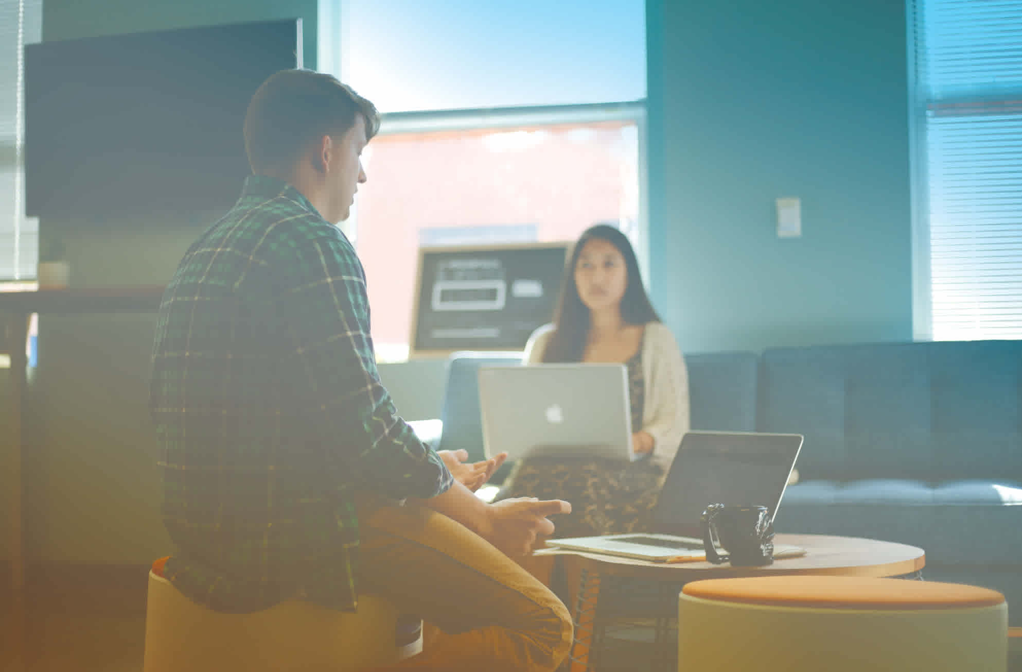 A man and woman talk while they work on laptops in an office.