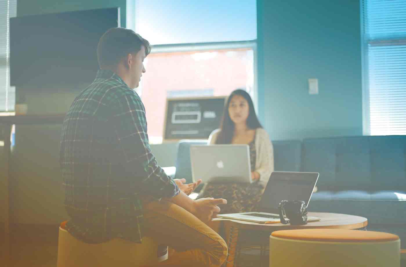 A man and woman talk while they work on laptops in an office.