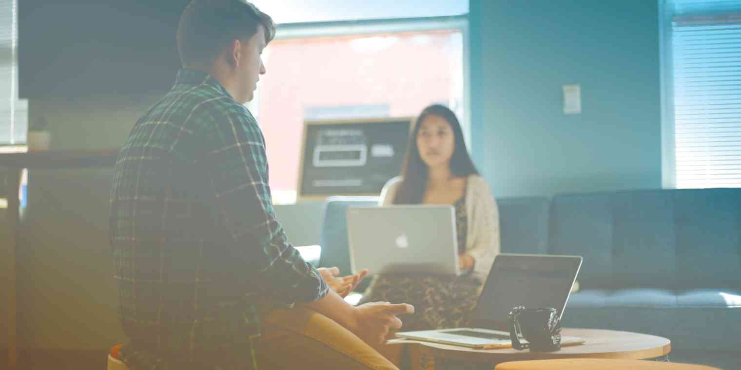 A man and woman talk while they work on laptops in an office.