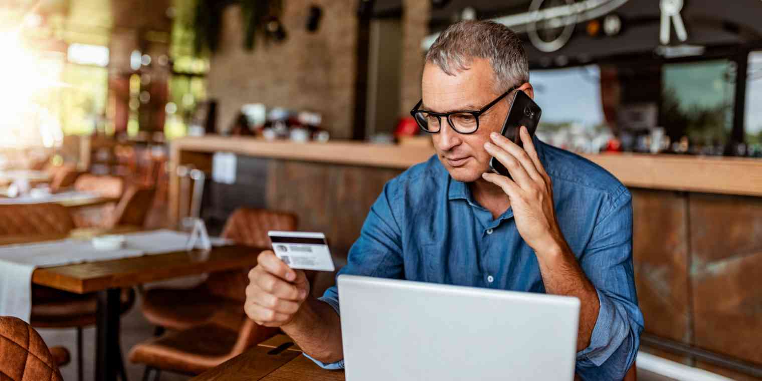 Hero image of a man at a coffee shop, holding a credit card while on the phone, with a computer in front of him