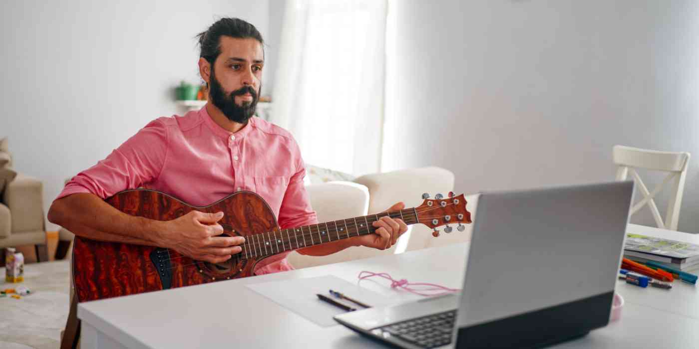 Hero image of a man playing guitar in front of a computer (virtual music lesson)
