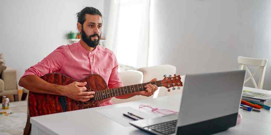 Hero image of a man playing guitar in front of a computer (virtual music lesson)