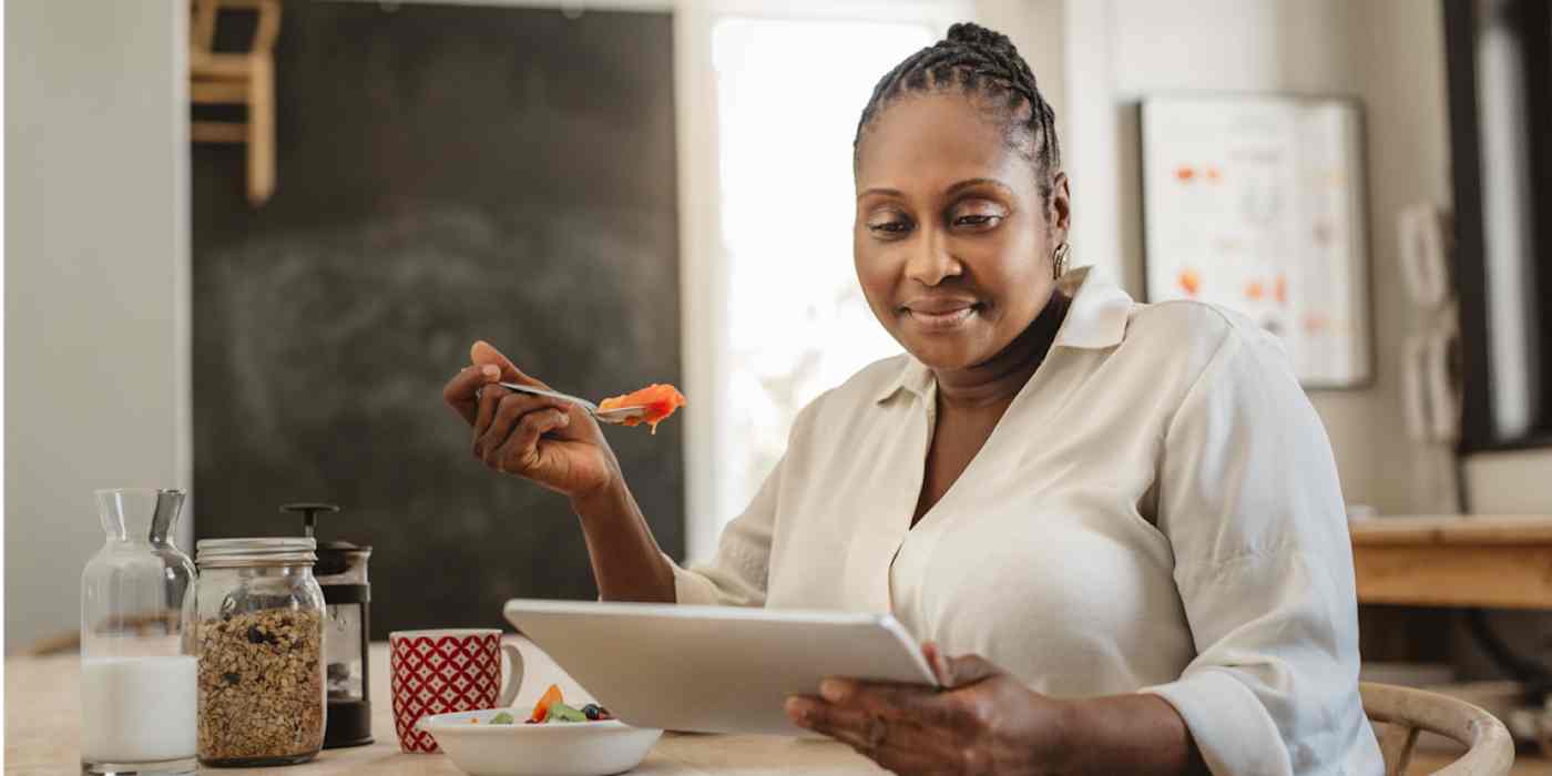 Woman sitting in front of her kitchen table in a brightly lit room. She's holding a forkful of food in one hand and looking down at a tablet being held in the other hand.