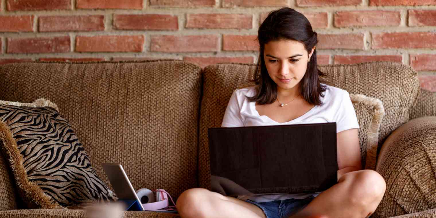 Hero image of a young woman sitting on a couch with a computer, with a brick wall behind her