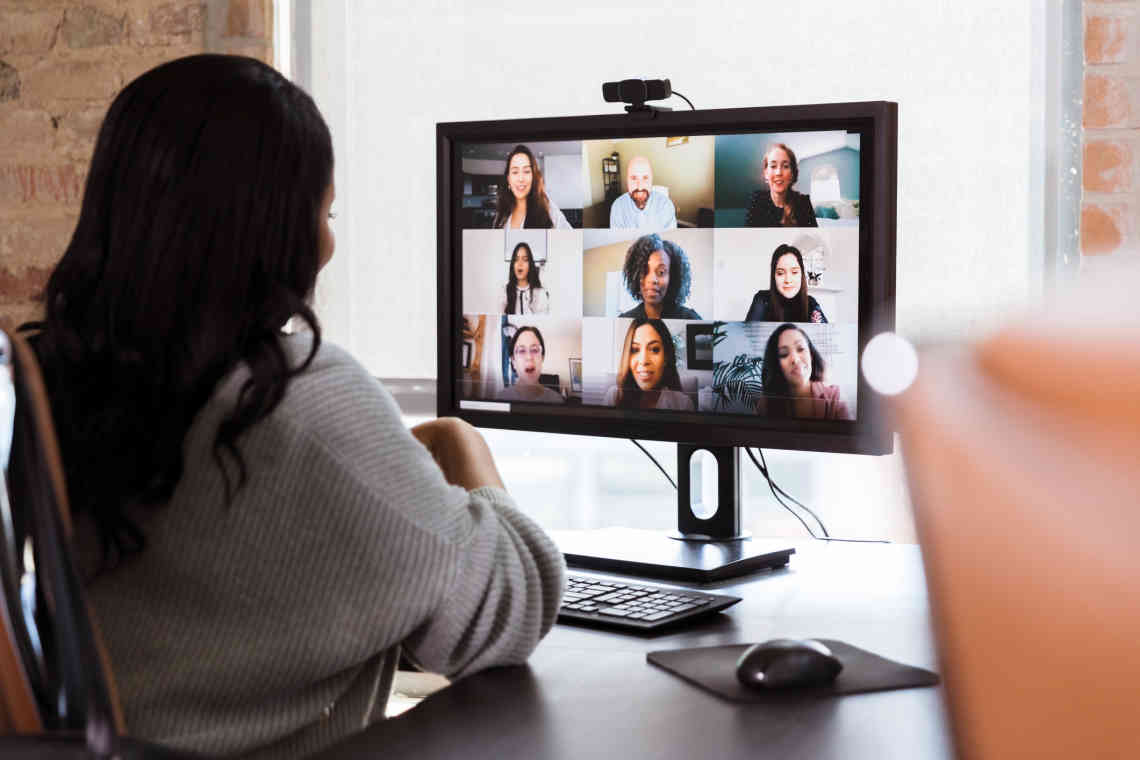 A woman sits at a desk looking at a screen showing nine participants in a video call.