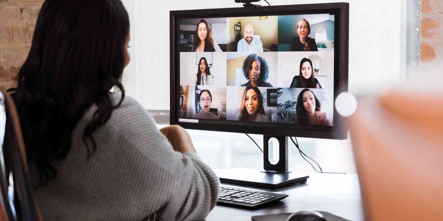 A woman sits at a desk looking at a screen showing nine participants in a video call.