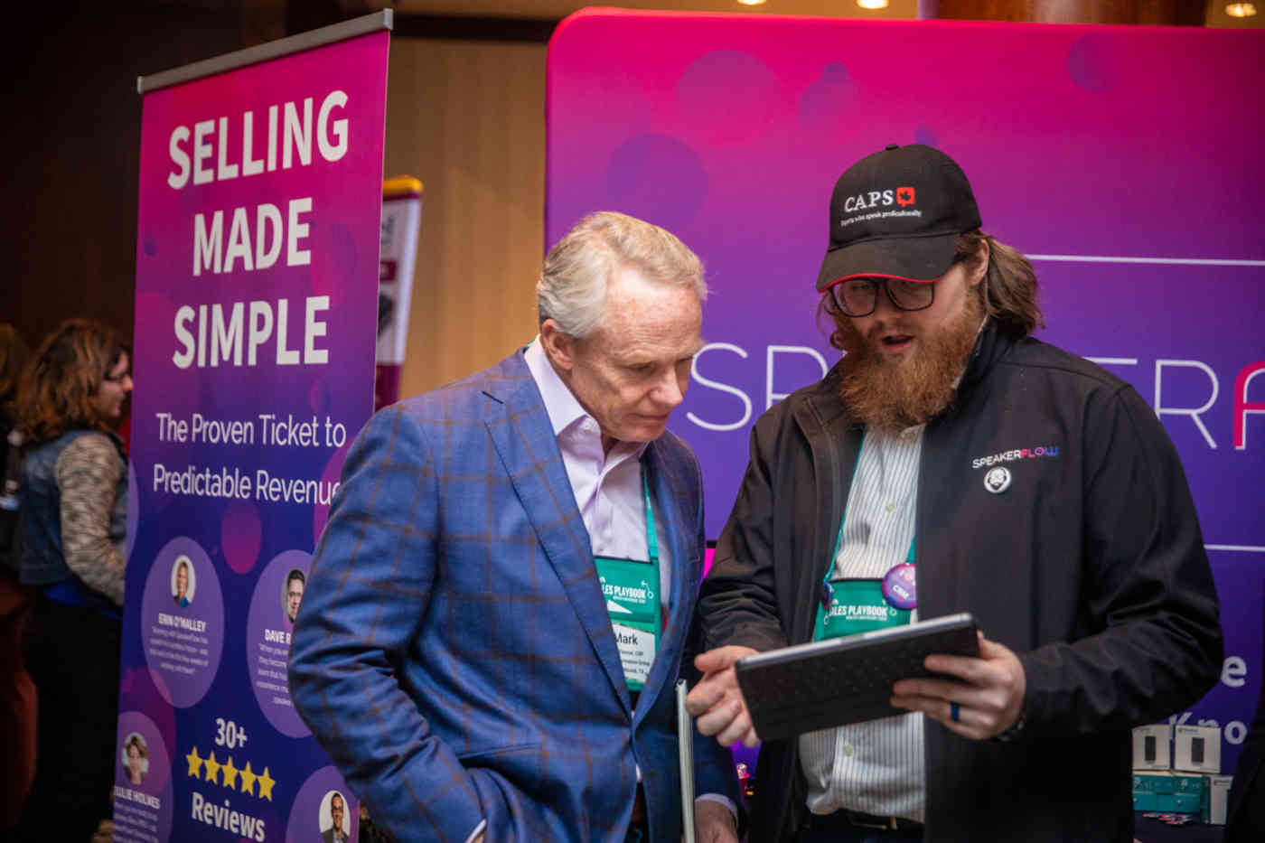 Two men stand in front of signage at a conference. One of them holds a tablet. The sign behind them reads "Selling made simple."