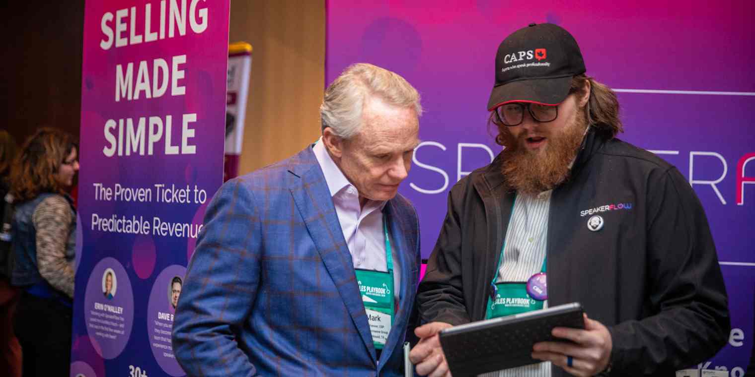 Two men stand in front of signage at a conference. One of them holds a tablet. The sign behind them reads "Selling made simple."