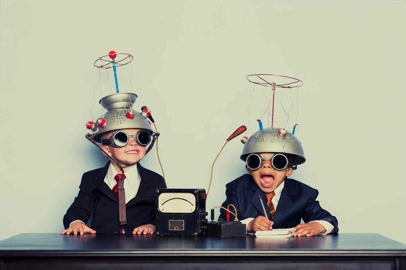 Two grinning children sit at a table wearing goggles with colanders on their heads.