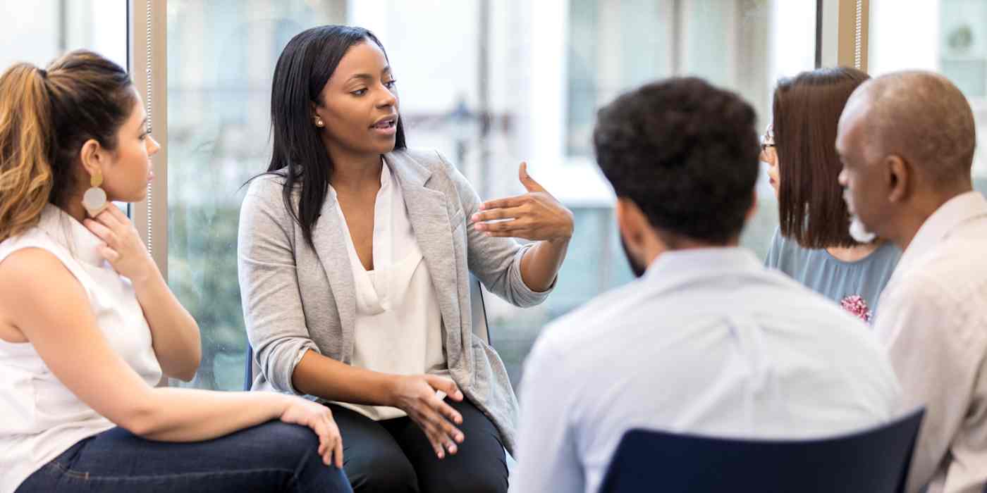 Hero image of a woman speaking to a small group of people, all seated in a circle