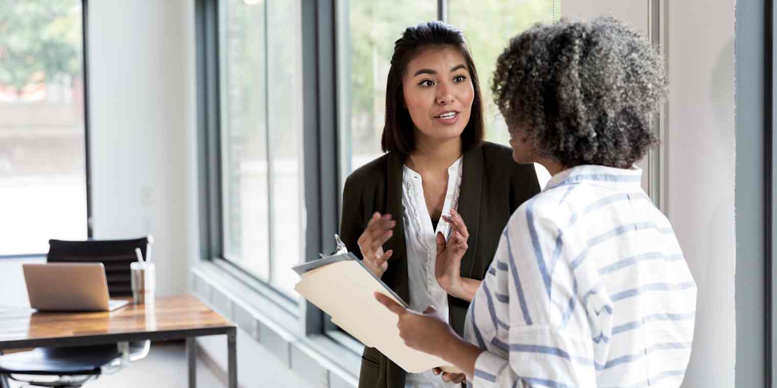 Hero image of two women talking. One of them is gesticulating and the other is holding a folder.