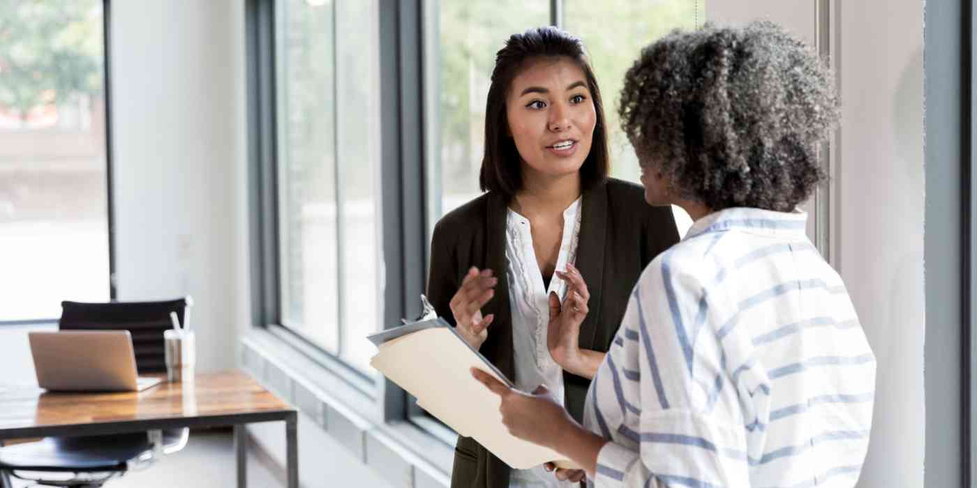 Hero image of two women talking. One of them is gesticulating and the other is holding a folder.