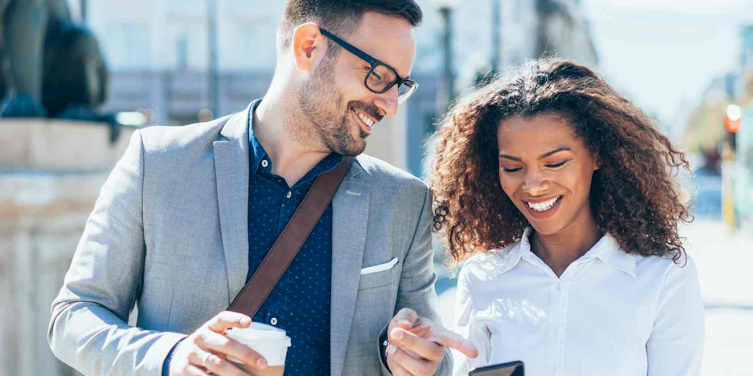 A man and woman walk down a city street. They are smiling and he is pointing at something on her phone screen.
