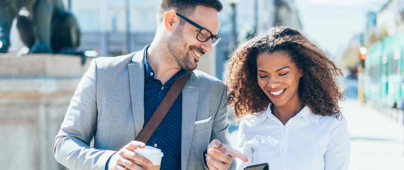 A man and woman walk down a city street. They are smiling and he is pointing at something on her phone screen.