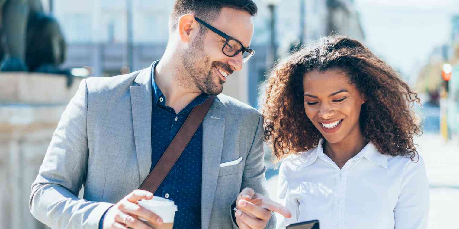 A man and woman walk down a city street. They are smiling and he is pointing at something on her phone screen.