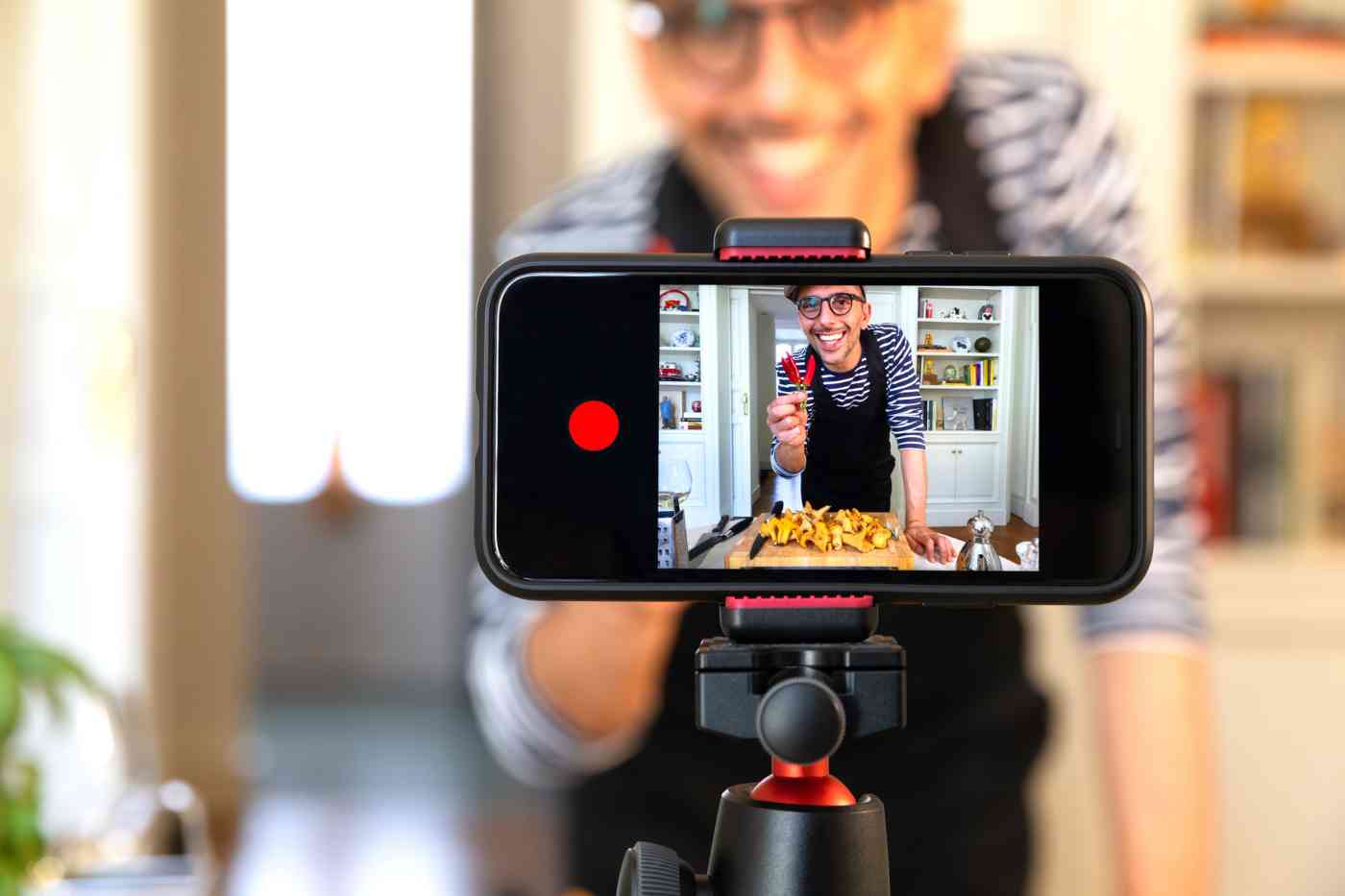 A phone held horizontally in a tripod displaying an image of a man holding several peppers. He is recording a video of himself cooking something.
