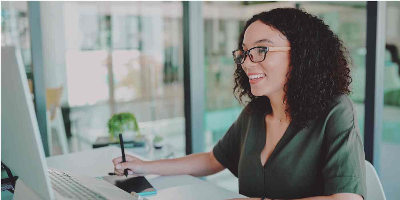 Hero image of a woman smiling, looking at a computer, with a pen in her hand