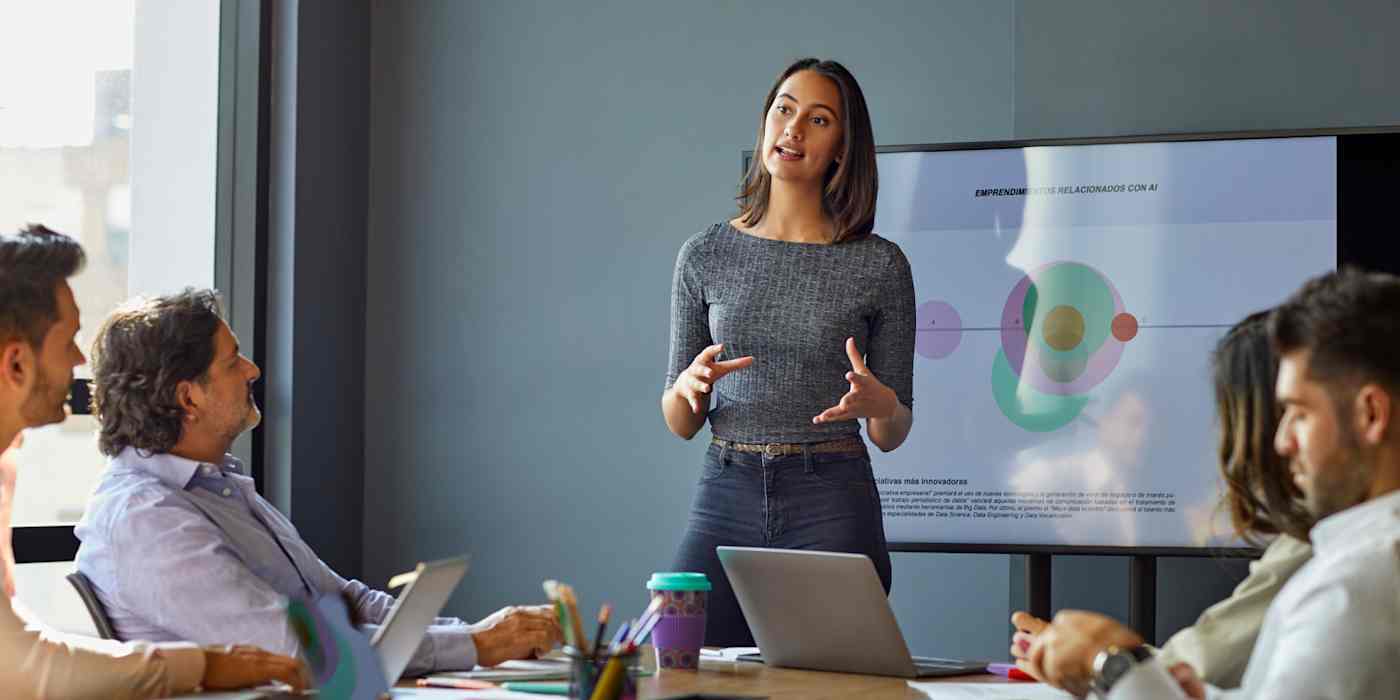 Hero image of a woman presenting to a conference room full of people, with a pie chart on a screen behind her