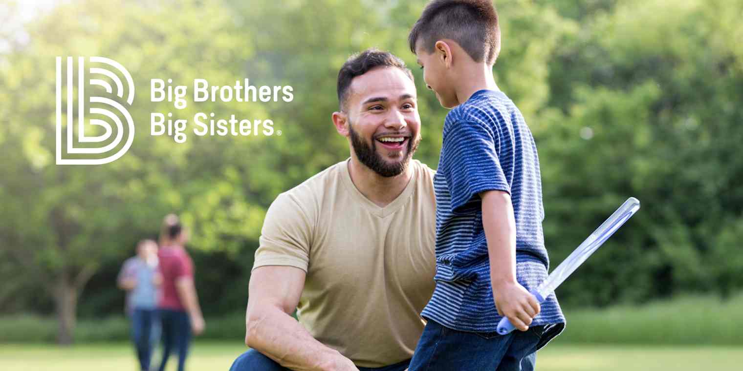 Outside in a field a smiling man kneels by a boy holding a large bubble wand