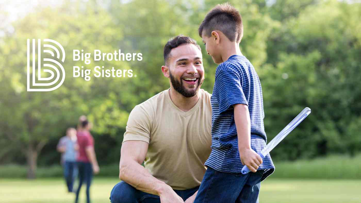 Outside in a field a smiling man kneels by a boy holding a large bubble wand