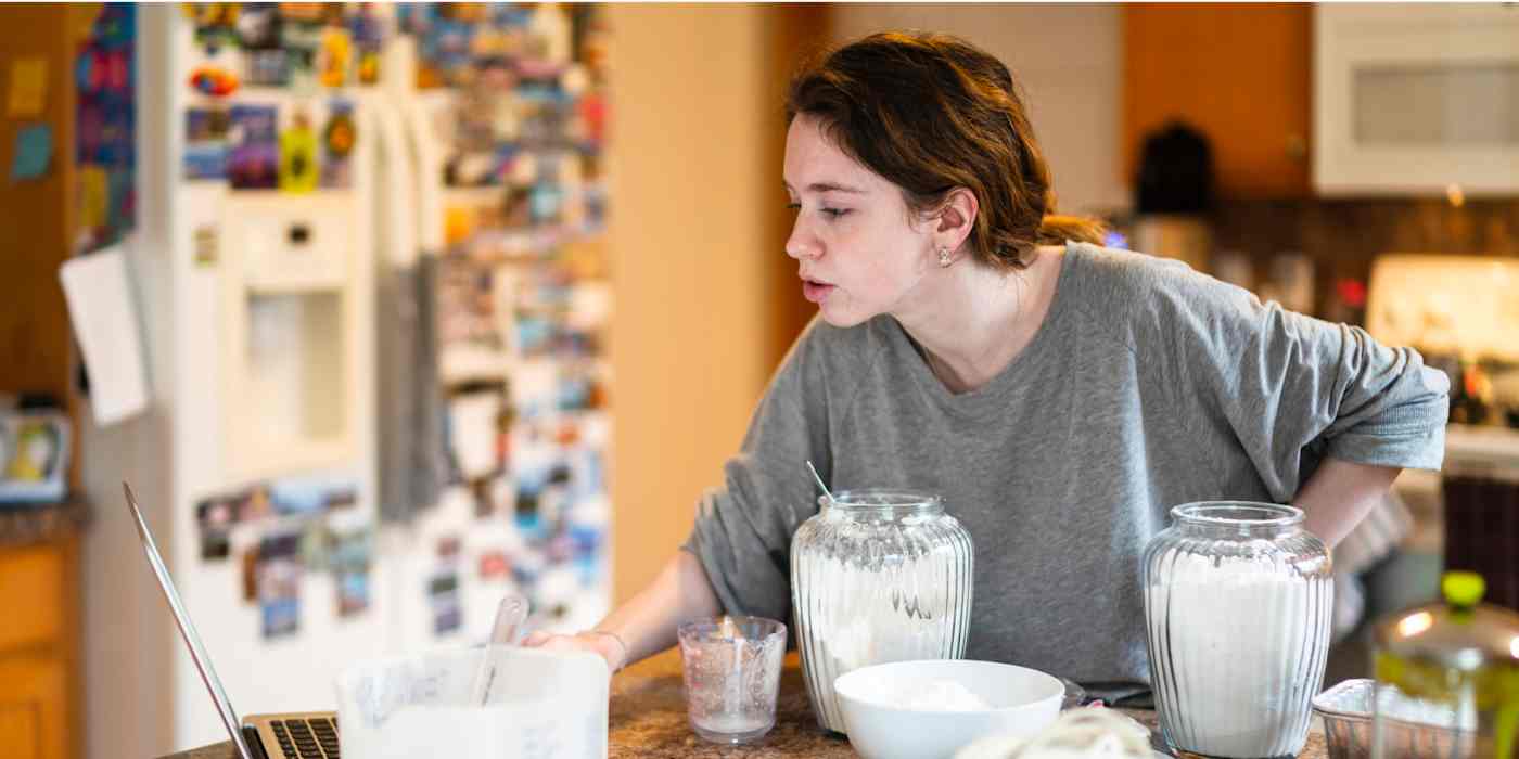 Hero image of a woman in a kitchen, baking and looking at a computer